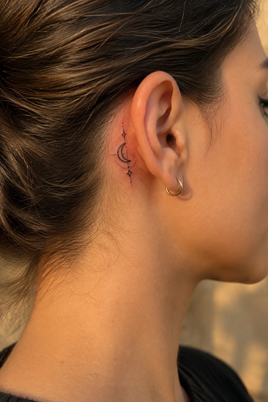 Small black crescent moon and star tattoo with dotted details behind a woman’s ear, above a gold hoop earring.
