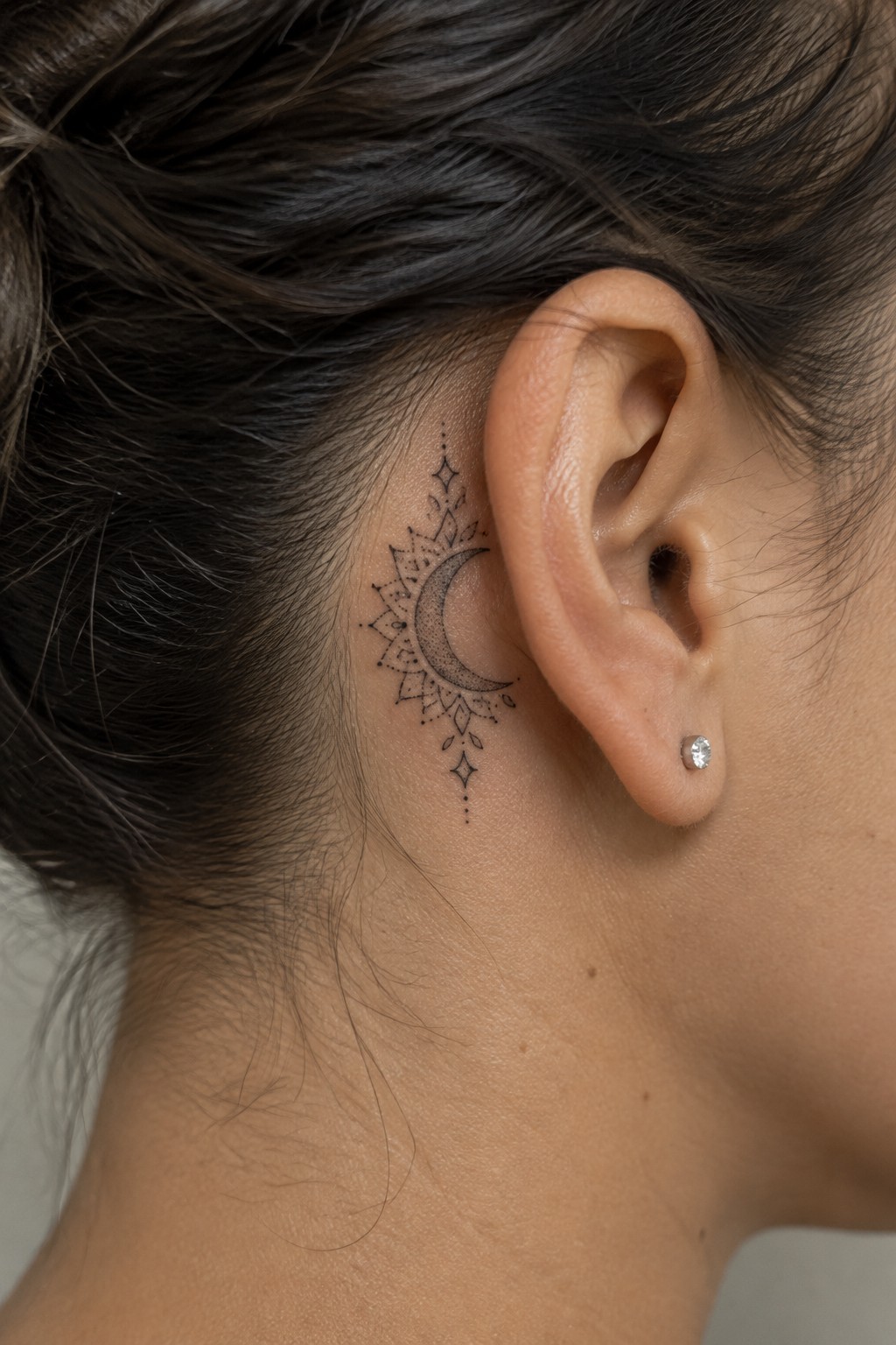 Small black crescent moon tattoo surrounded by fine-line mandala details behind a woman’s ear.