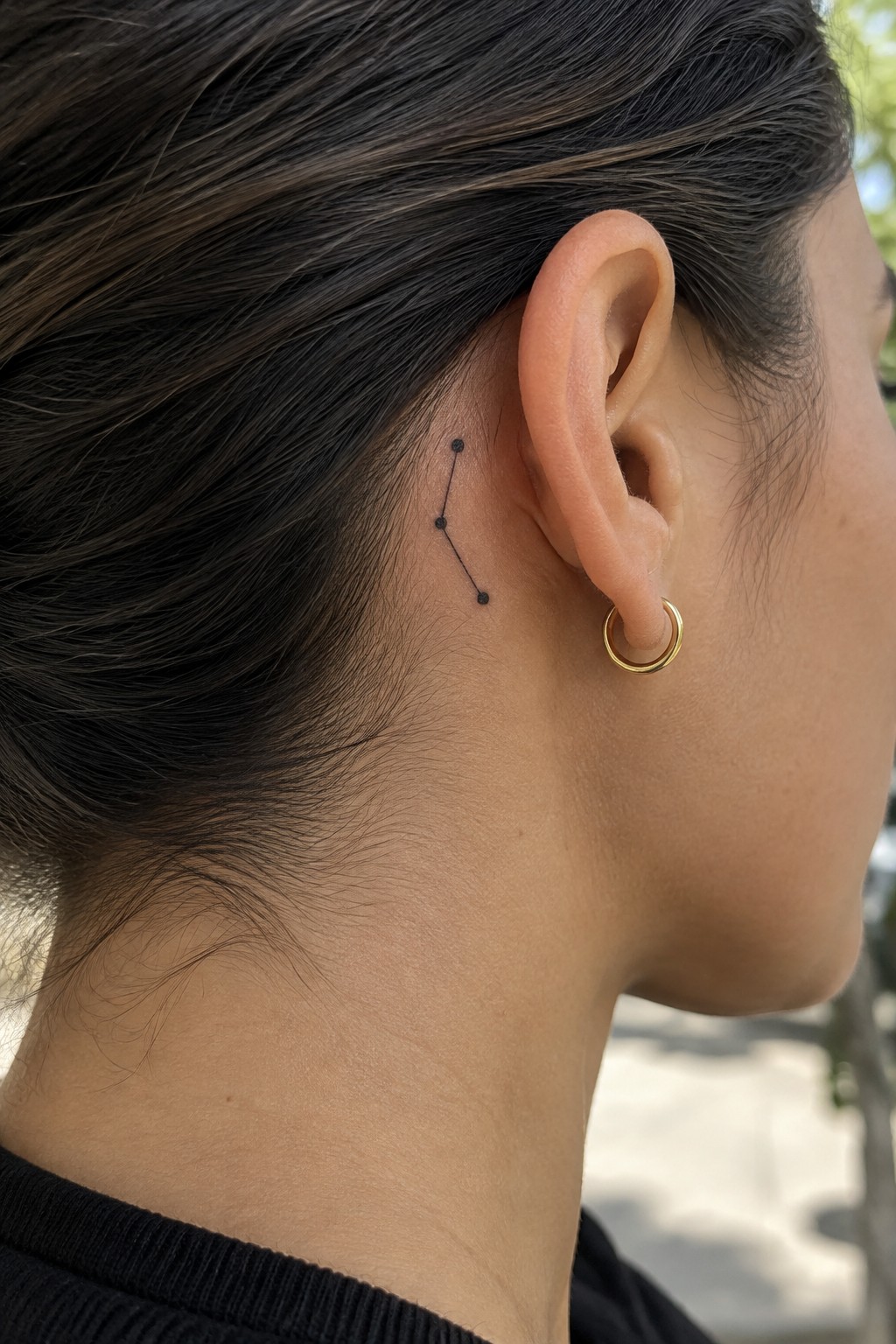 Small black constellation tattoo with three dots and fine connecting lines behind a woman’s ear.