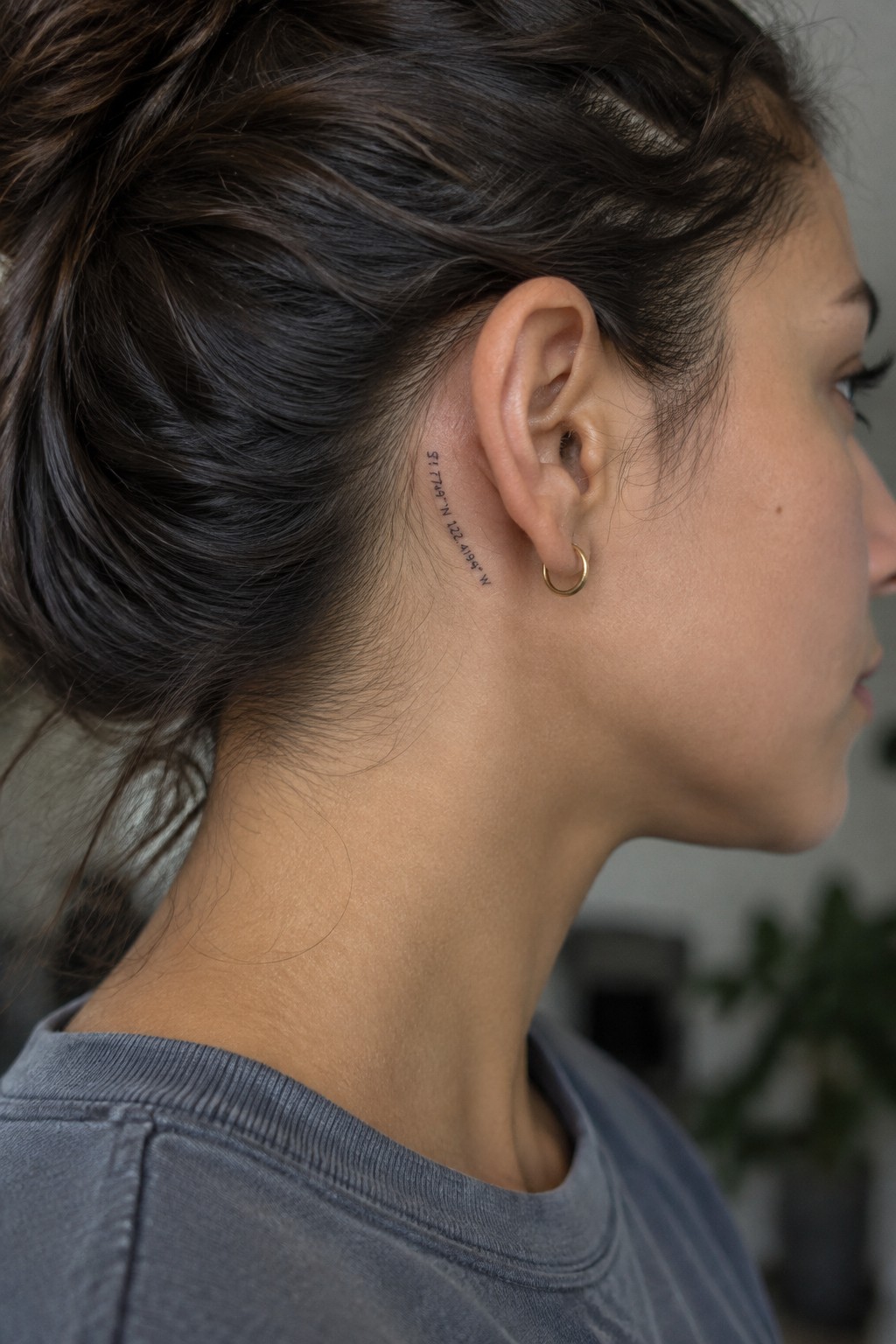 Fine-line geographic coordinates tattoo behind a woman’s ear, beside a small gold hoop earring.