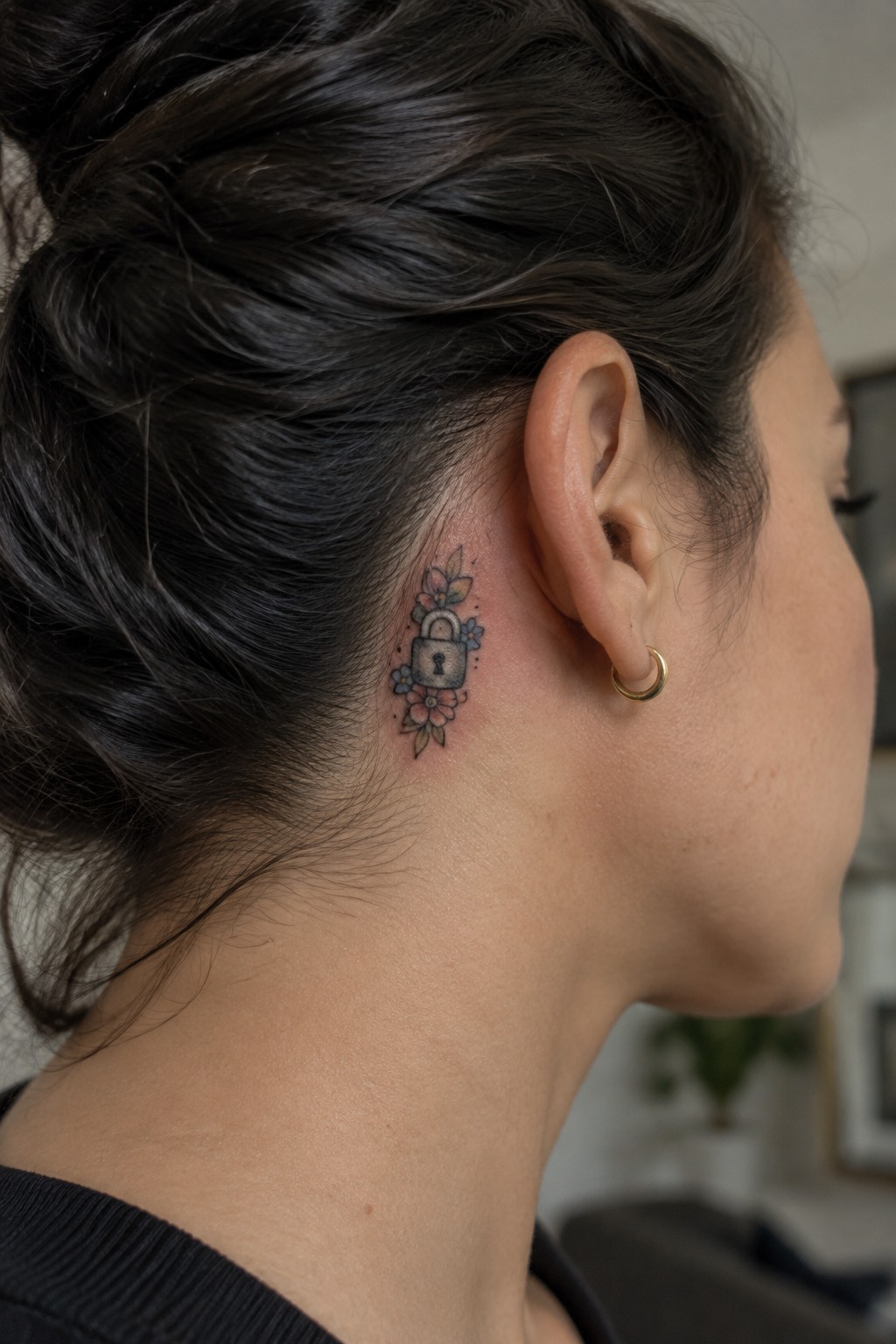 Small colored padlock tattoo surrounded by flowers behind a woman’s ear.
