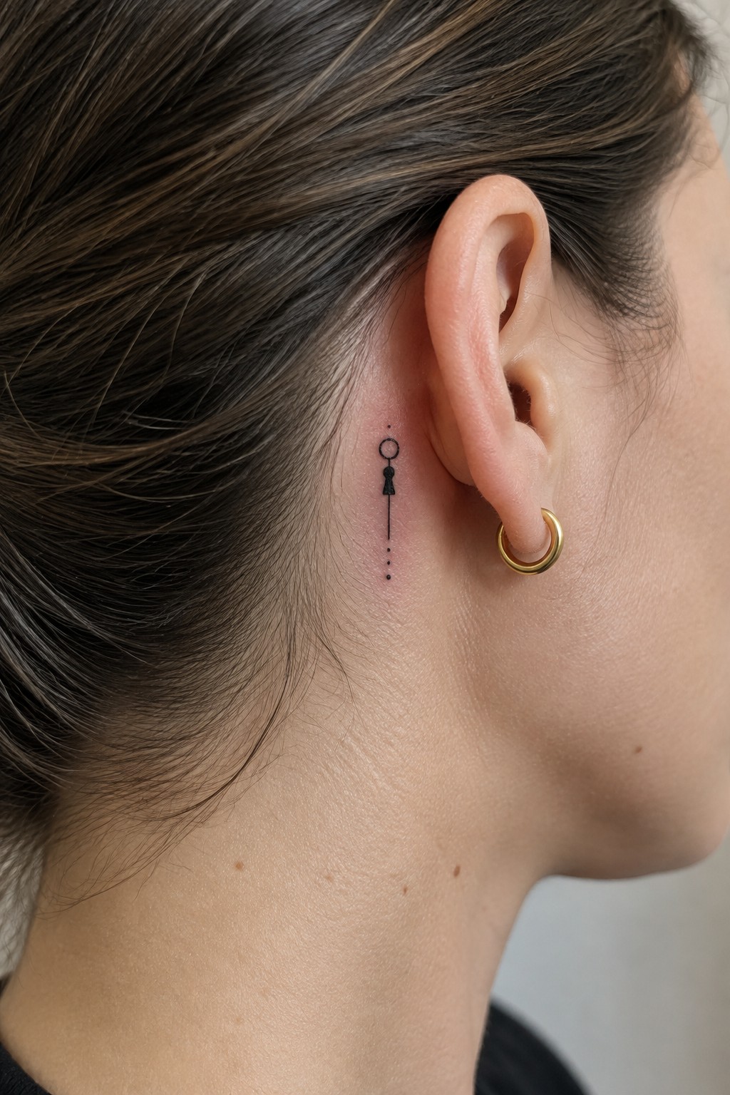 Small black geometric tattoo with a circle, solid shape, vertical line, and dots behind a woman’s ear, next to a gold hoop earring.