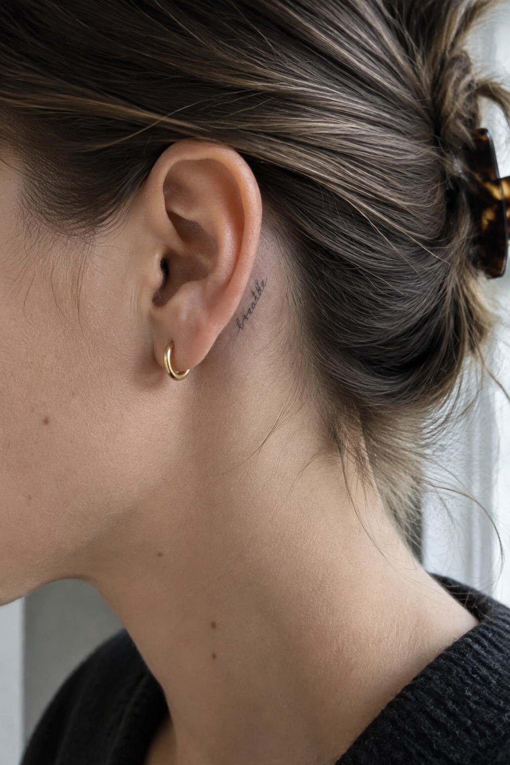 Small black script tattoo behind a woman’s ear, beside a gold hoop earring.
