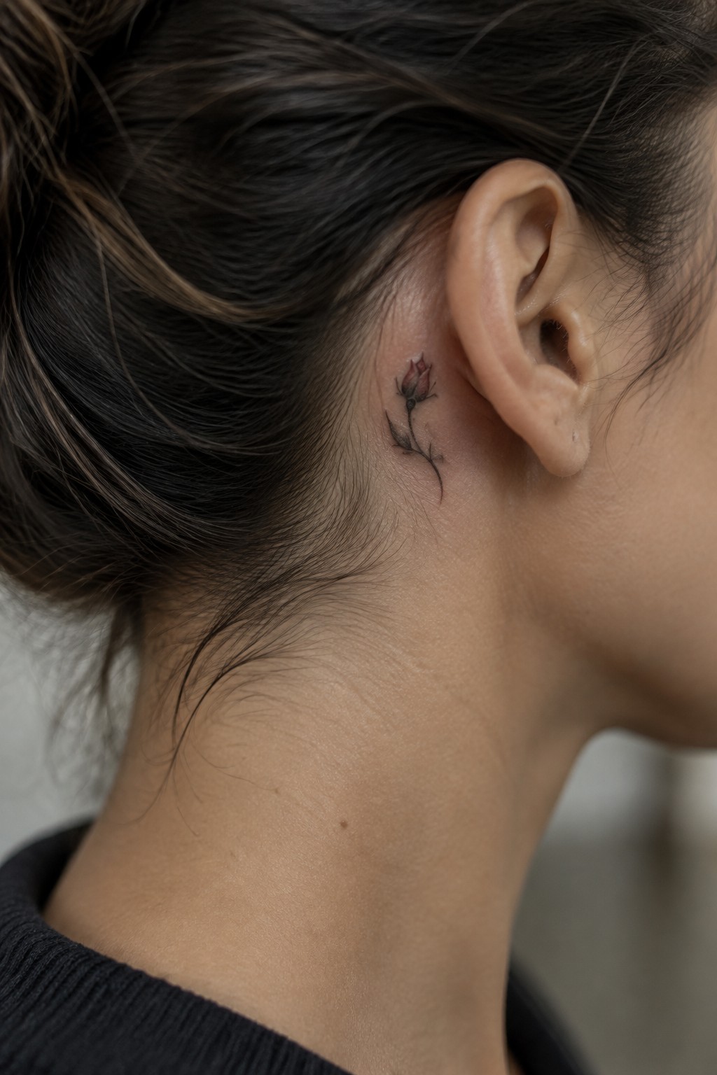 Small rosebud tattoo with a curved fine-line stem behind a woman's ear.