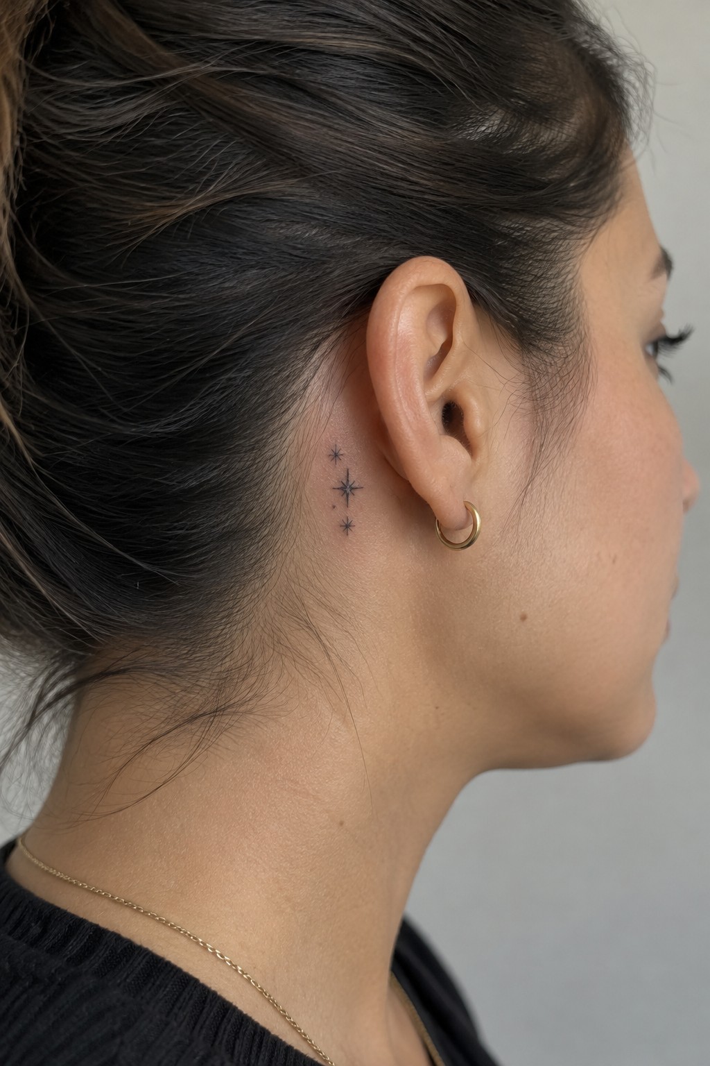 Three small black fine-line star tattoos in a vertical row behind a woman’s ear, beside a gold hoop earring.