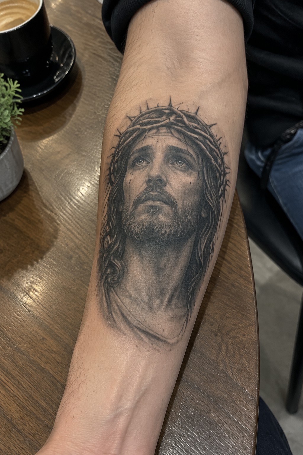 Realistic black-and-gray Jesus tattoo with crown of thorns on forearm beside coffee cup