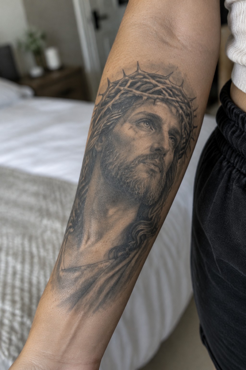 Realistic black and grey Jesus Christ tattoo with crown of thorns on the inner forearm, showing detailed shading and a vertical portrait layout.
