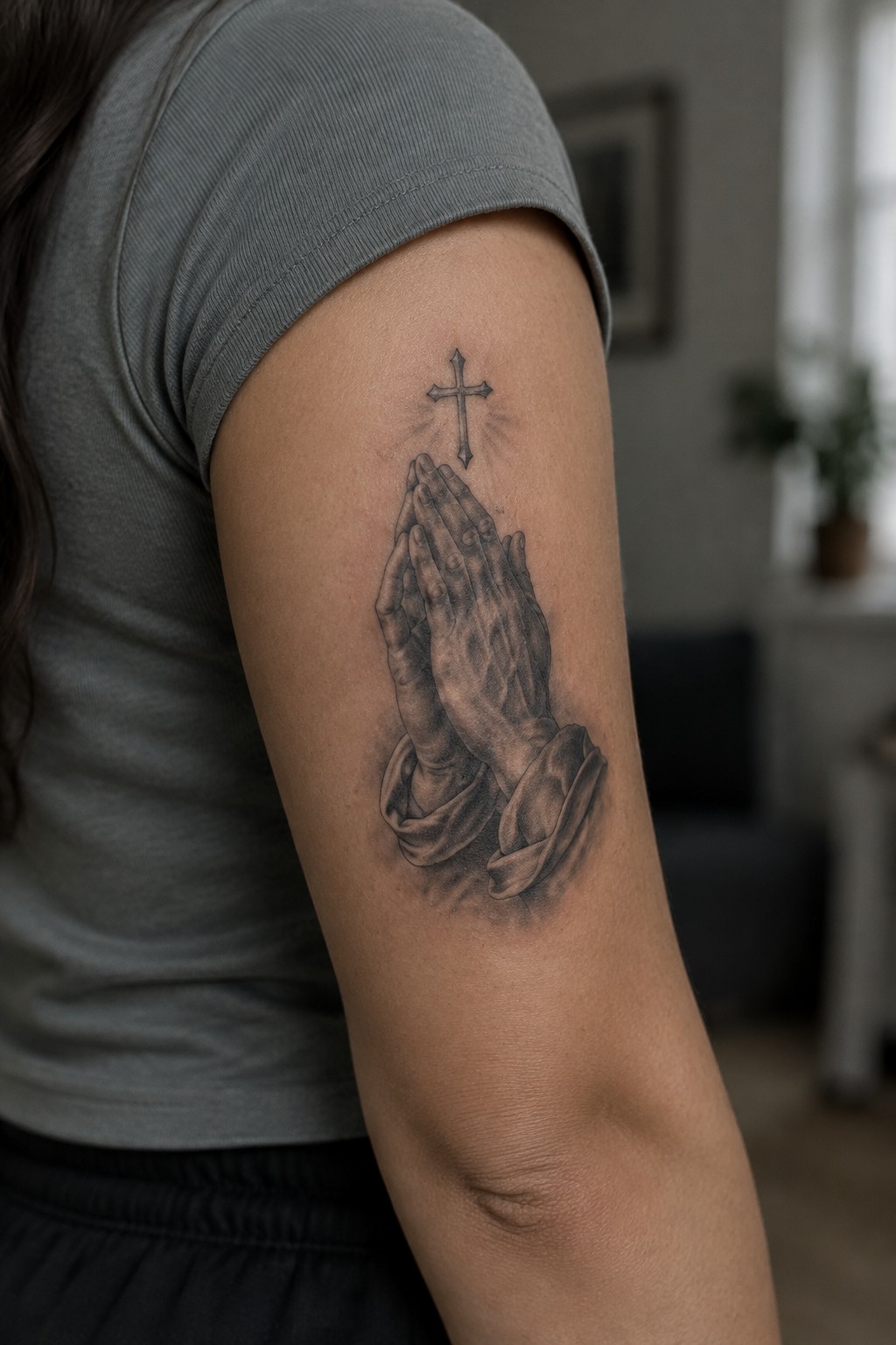 Black and gray tattoo of praying hands beneath a small cross on a woman’s upper arm.