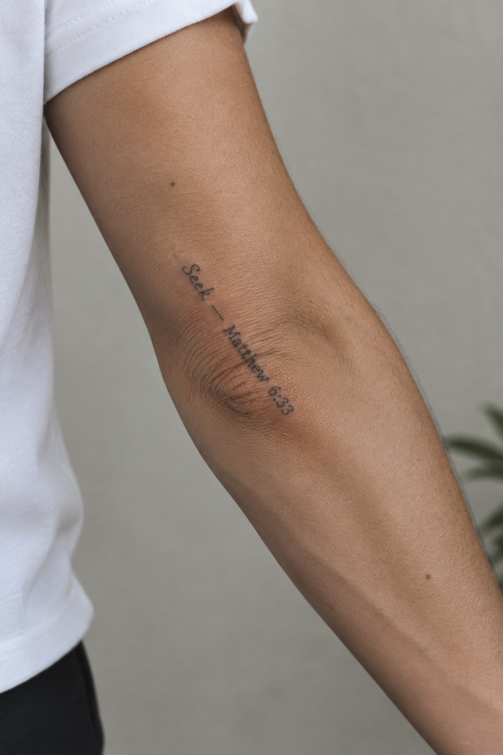 Close-up of arm with 'Seek — Matthew 6:33' tattoo beside white t-shirt sleeve.
