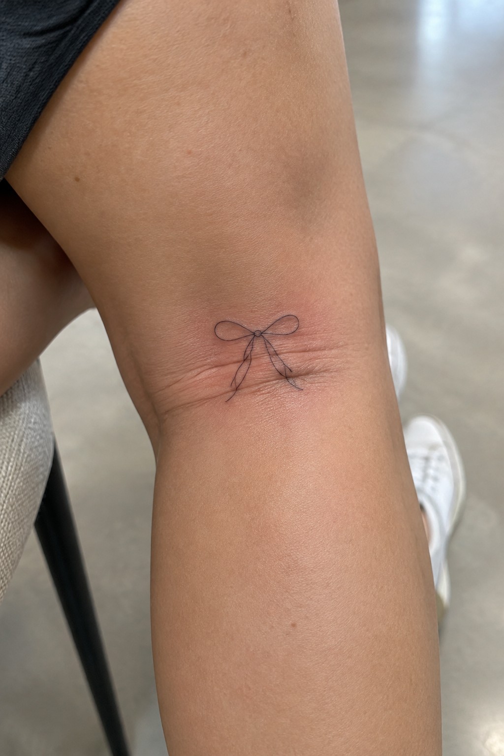Small black bow tattoo on fair skin, dark shorts and white sneaker visible.