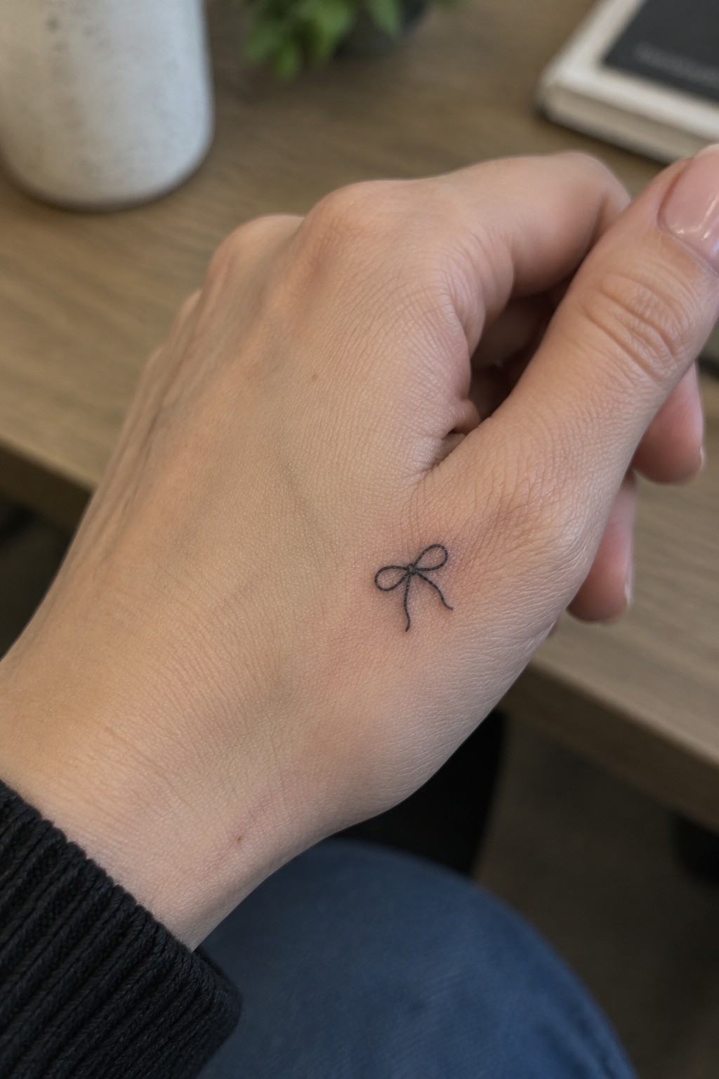 Hand with small black bow tattoo on wooden table, black sweater sleeve visible