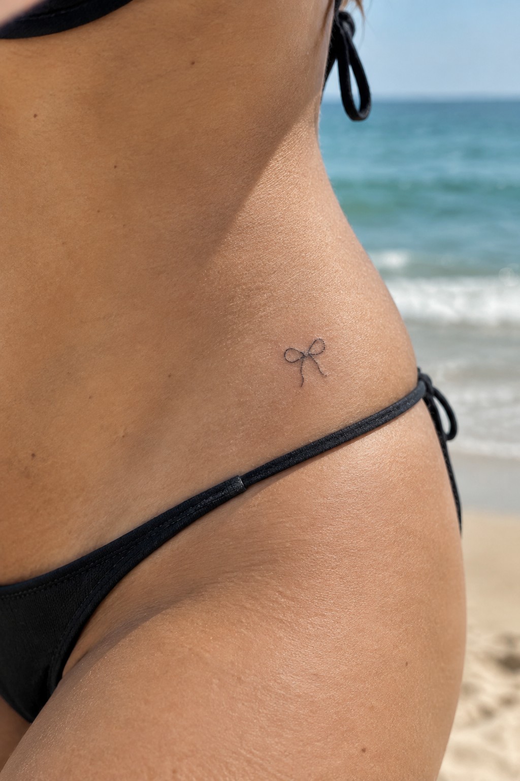 Small black bow tattoo on hip above black bikini bottoms at sunny beach