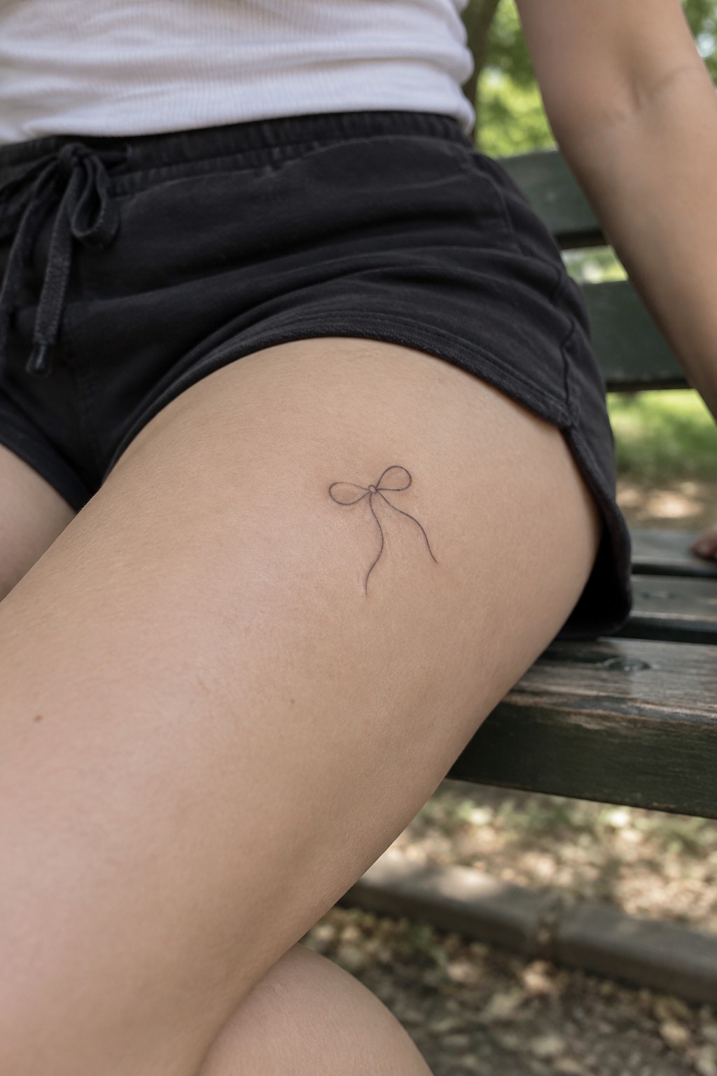 Small bow tattoo on thigh of person in white shirt and black shorts on bench