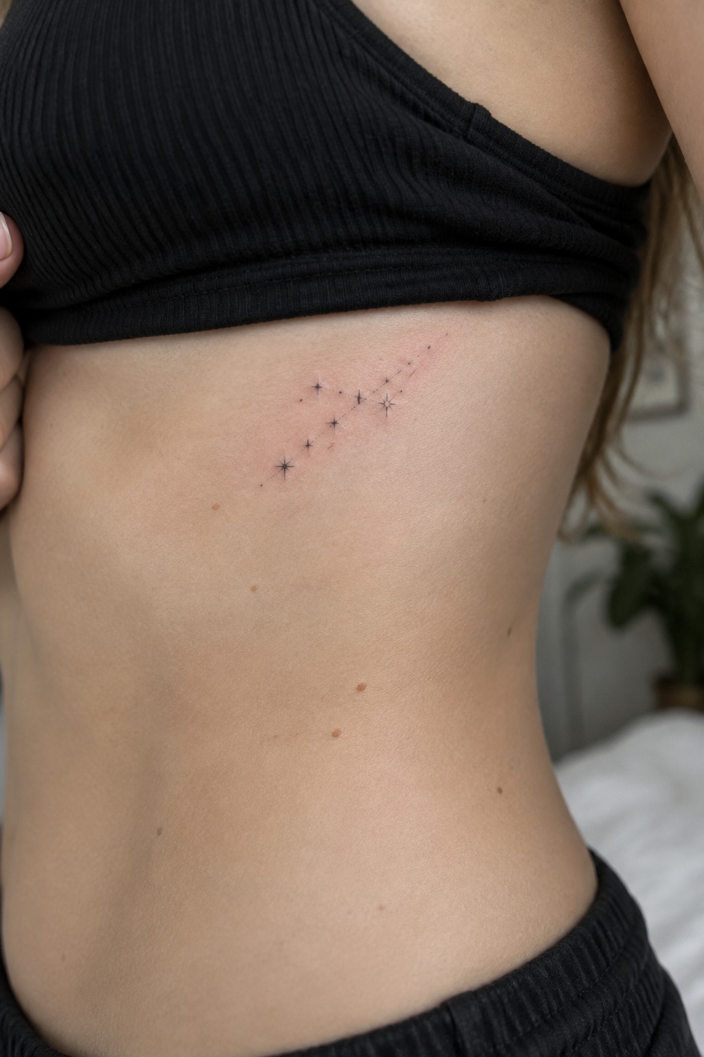 Diagonal star constellation tattoo on skin under lifted black crop top.