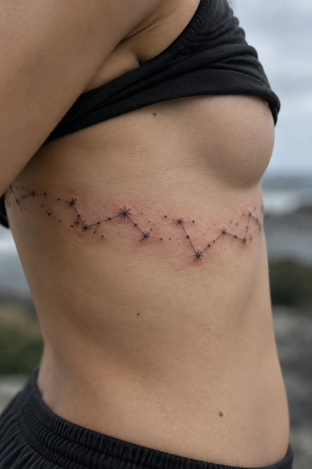 Constellation tattoo across bare side torso beneath lifted black top, skin reddened.