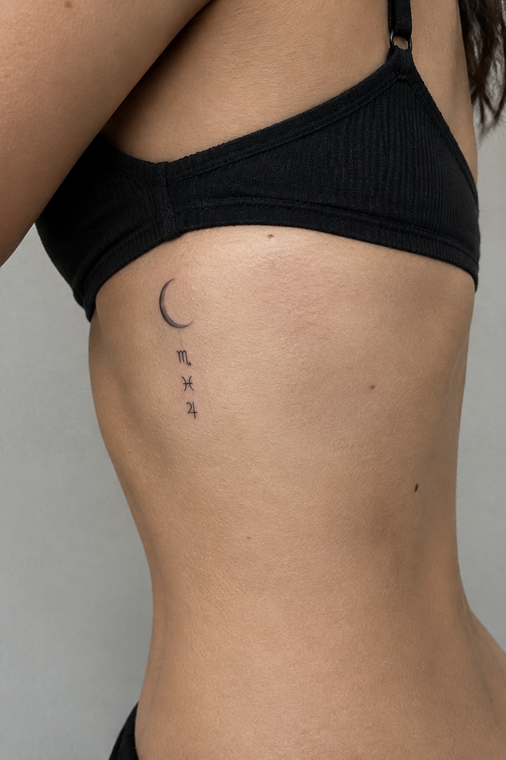 minimalist crescent moon tattoo above vertical pisces symbol and small text on the ribs
