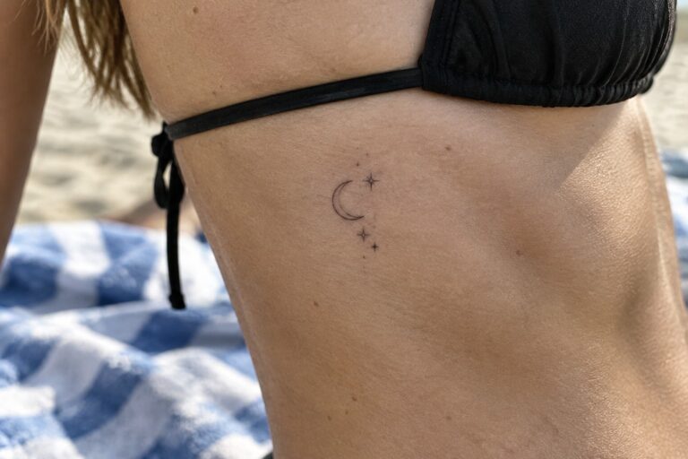 19 Minimalist Rib Tattoos Featuring Subtle Celestial Designs Black bikini strap on shoulder with crescent moon and stars tattoo at beach
