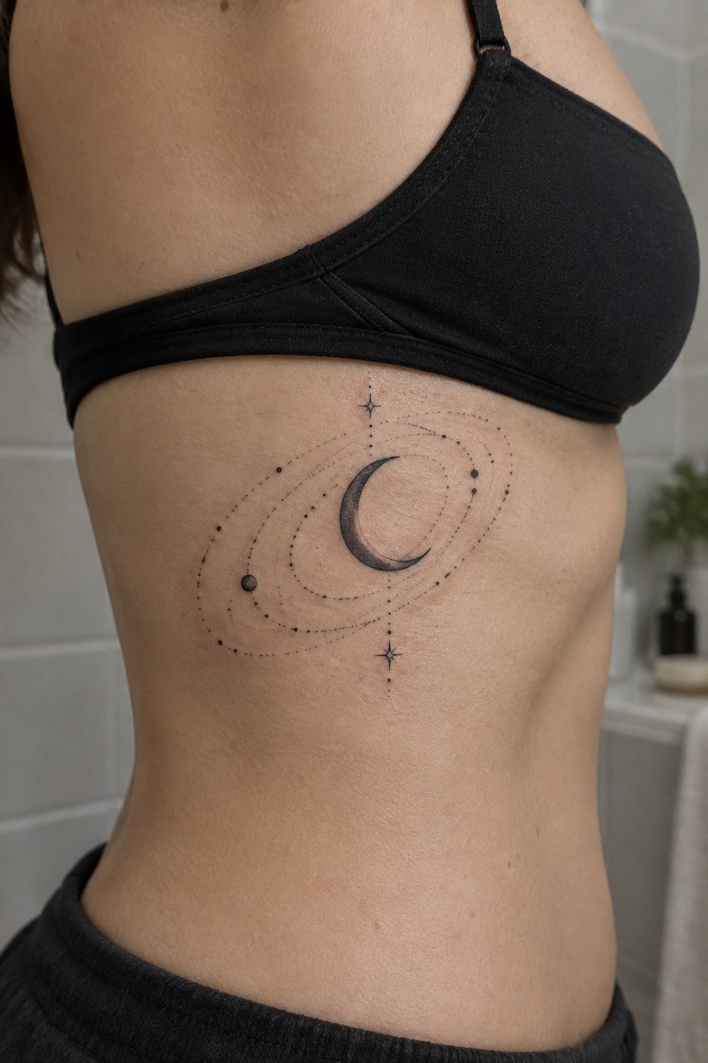 Minimalist rib tattoo with a crescent moon, dotted orbital rings, and small stars on the side torso.