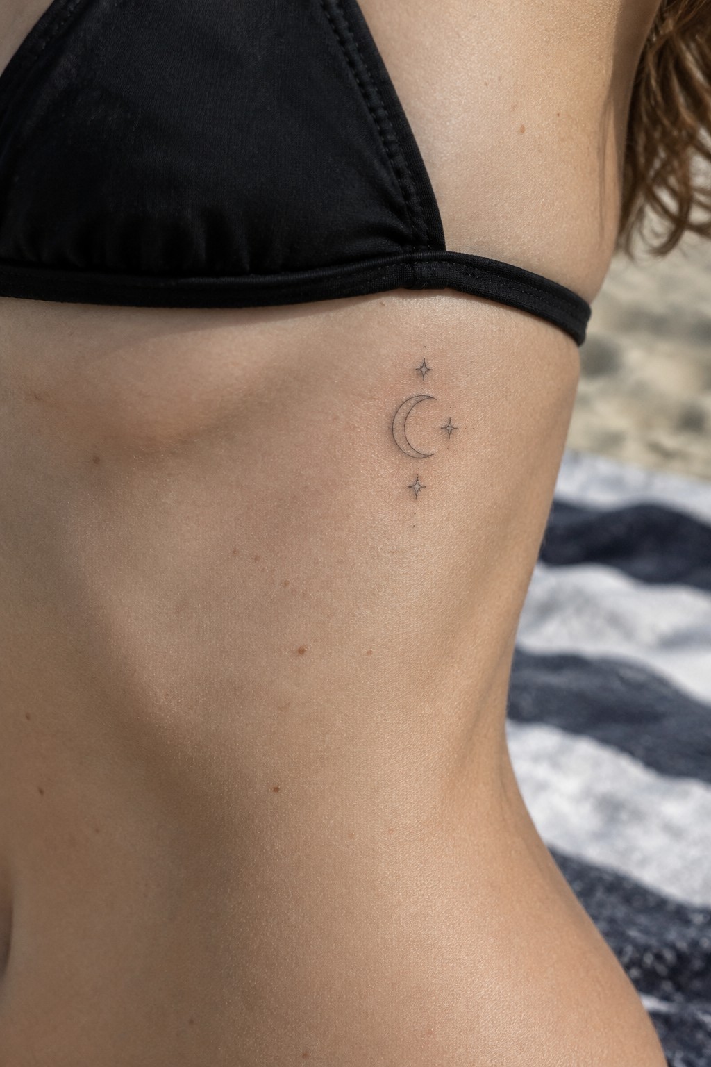 Black bikini strap on shoulder with crescent moon and stars tattoo at beach