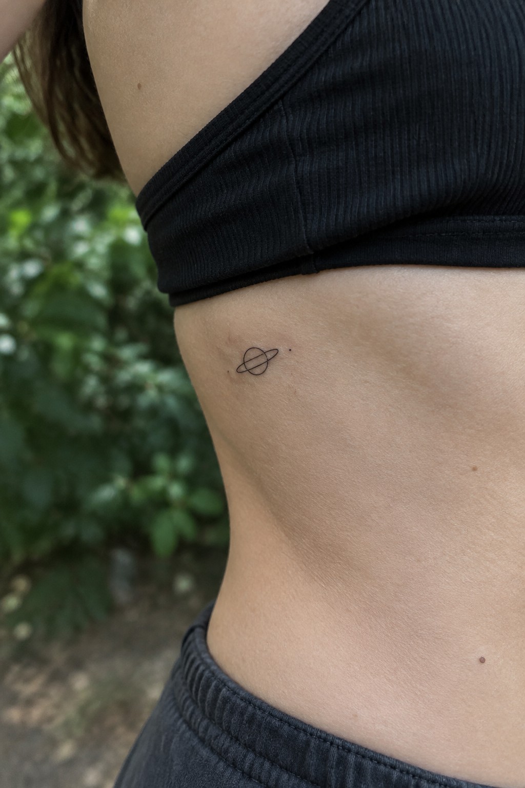 Small Saturn tattoo on bare skin beside black crop top and gray waistband.