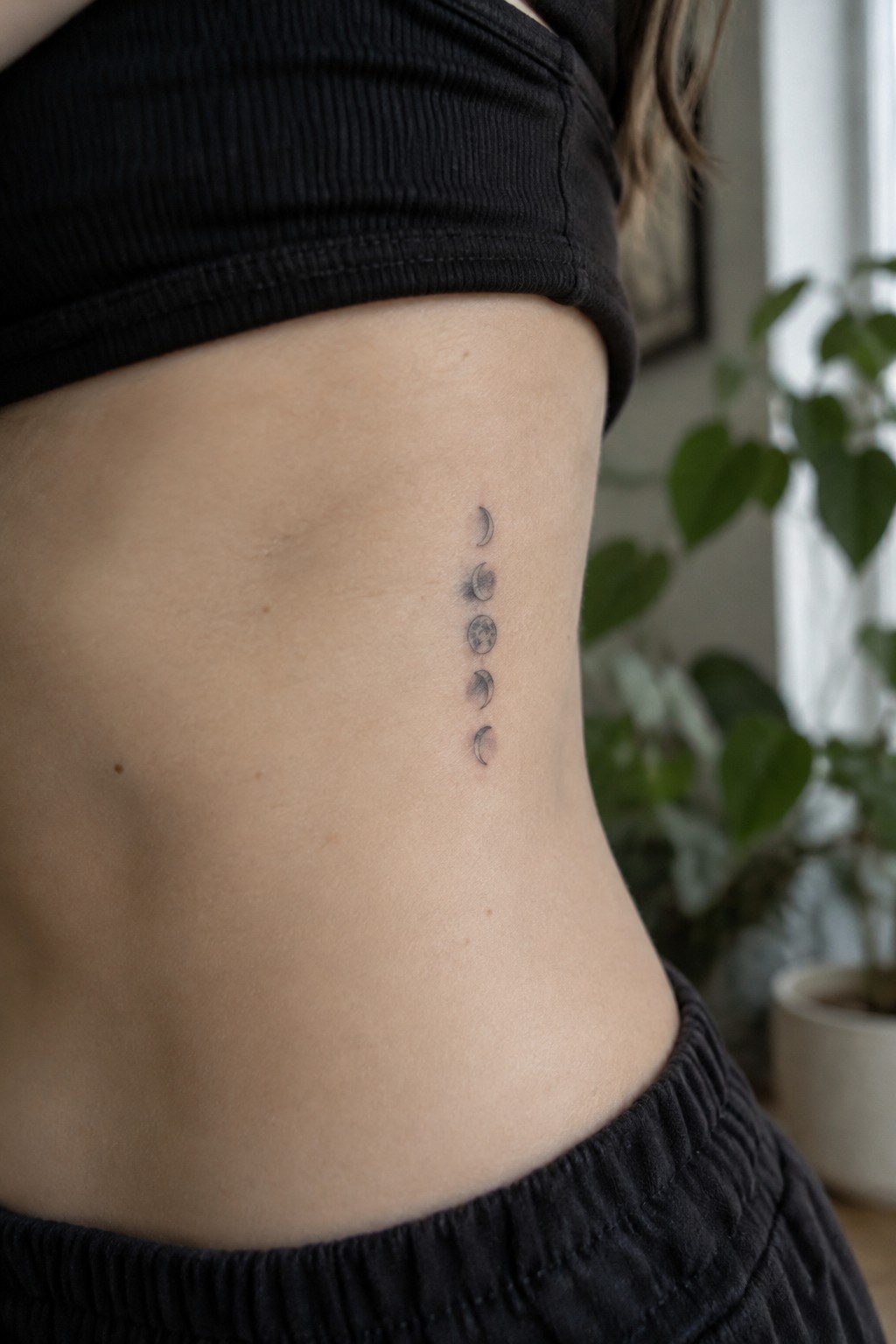 Moon phase tattoo on side under black crop top, blurred green plant behind.