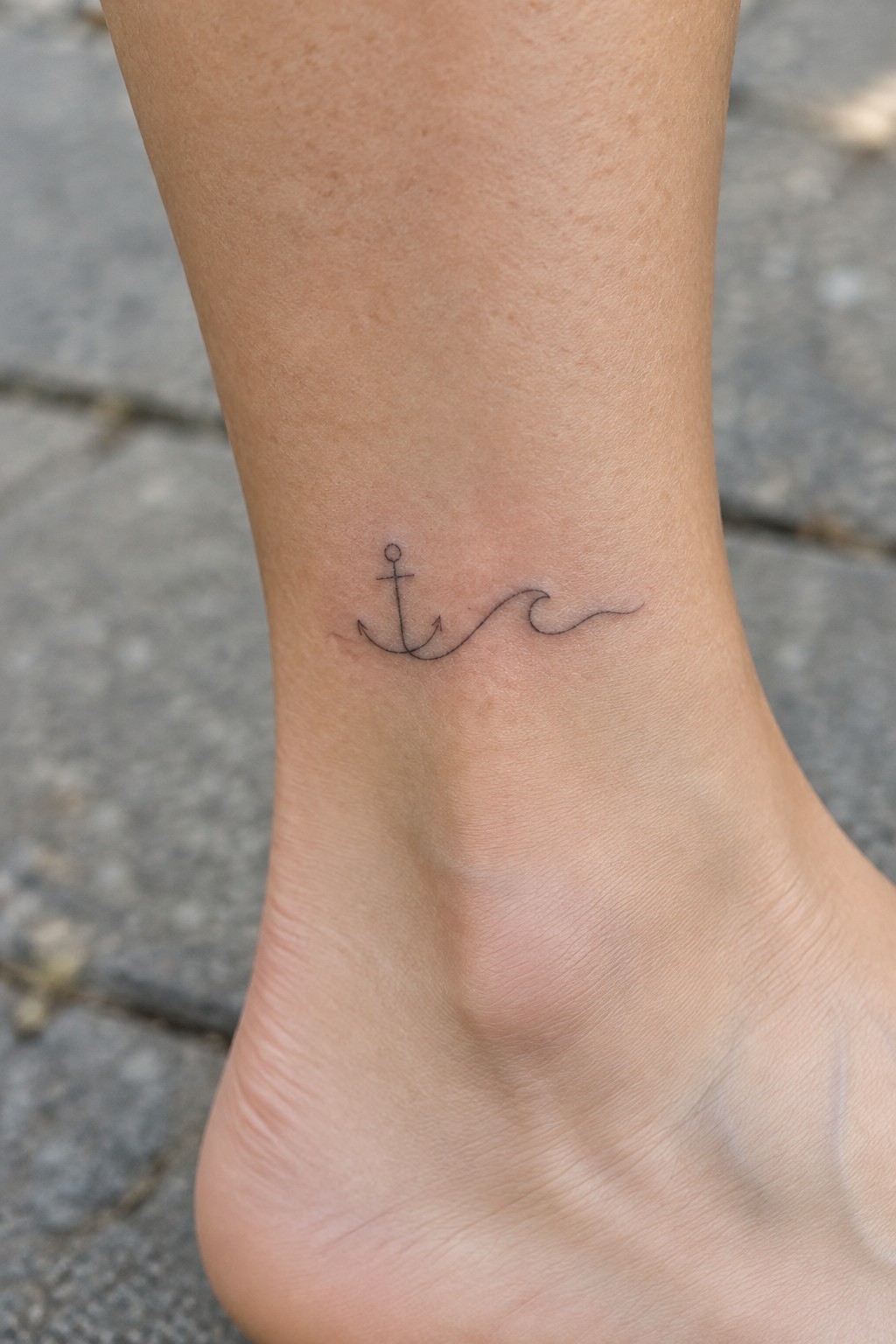 Minimalist black anchor and wave tattoo on bare ankle over pavement