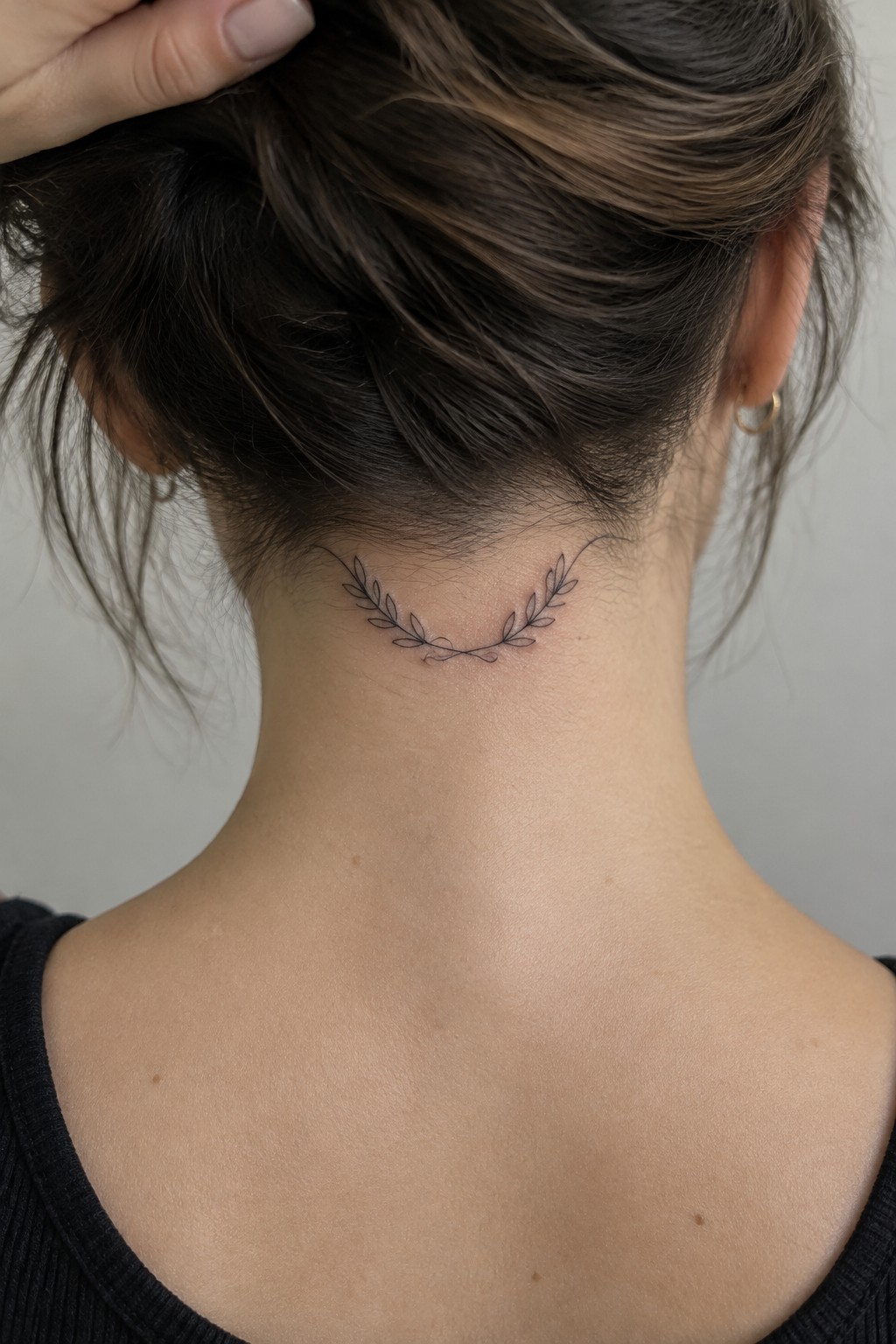 Thin continuous line laurel wreath tattoo curved across the back of a woman's neck below the hairline