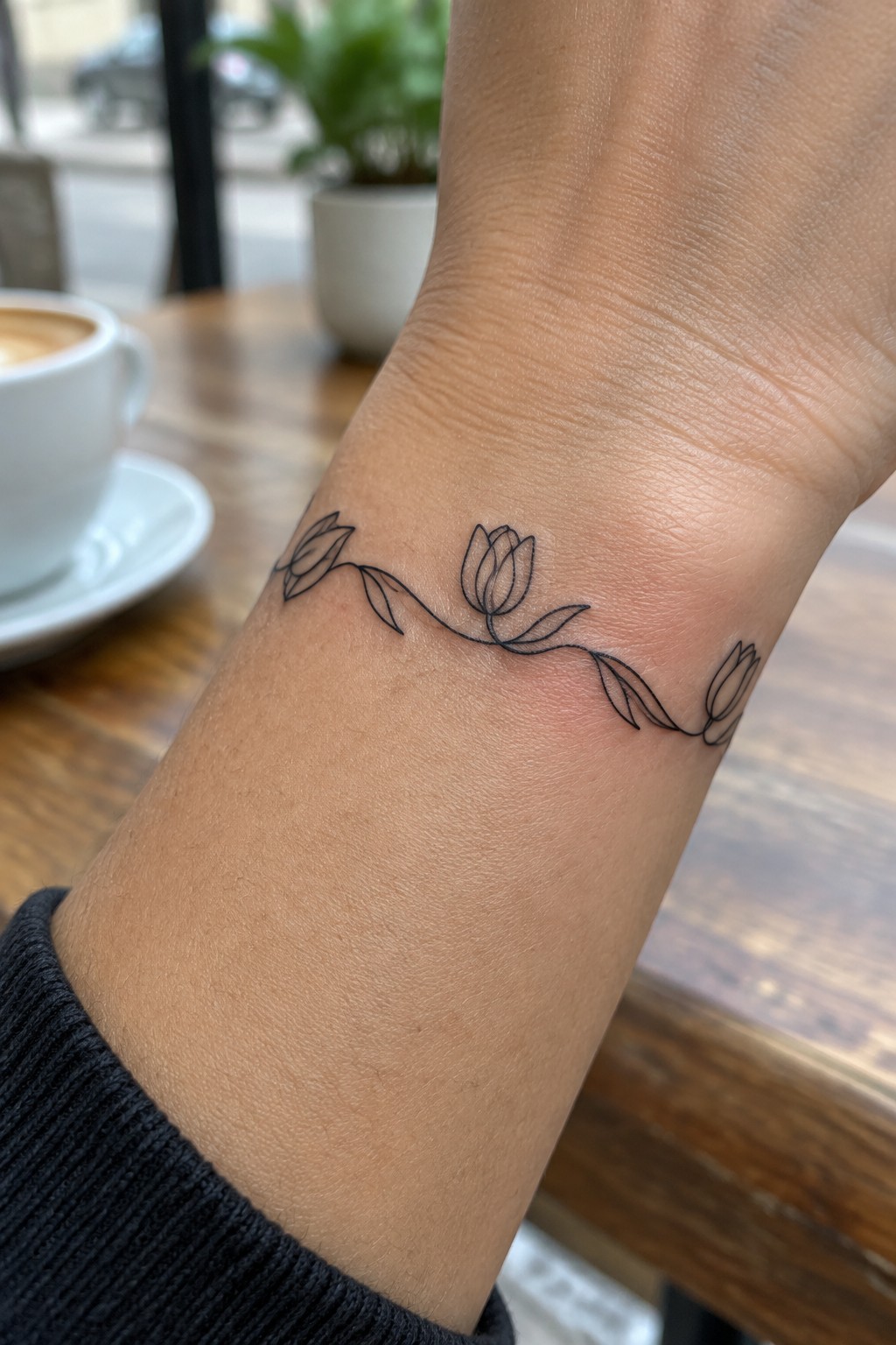 Wrist with delicate black line-art tulip vine tattoo near coffee cup
