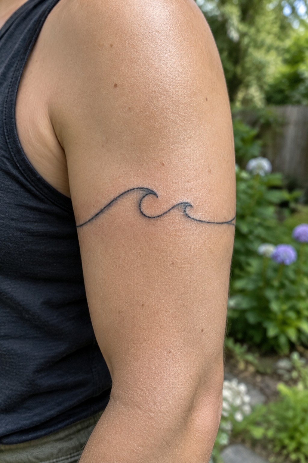 Wave tattoo on upper arm of person in black tank top outdoors.