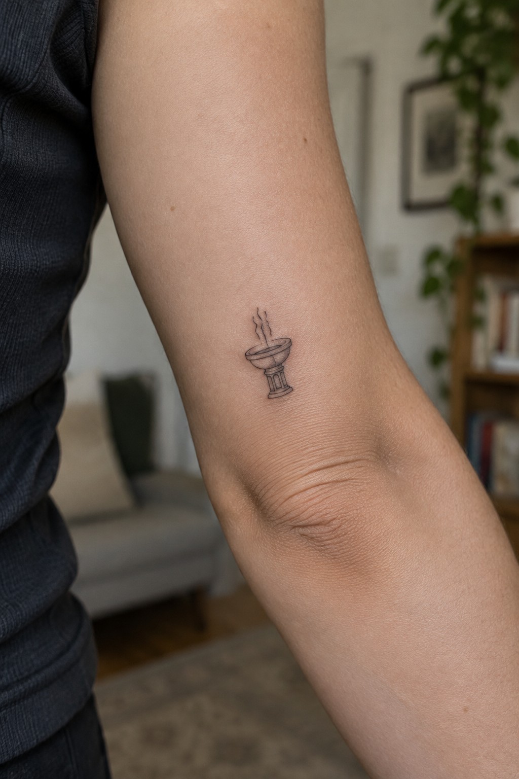 Small black ink smoking chalice tattoo on an arm beside a dark shirt.