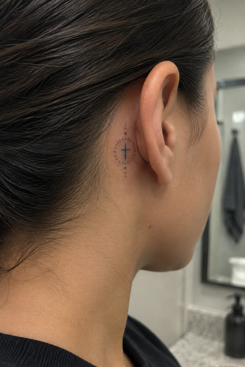 Dark-haired person displays small cross tattoo with dotted circle behind ear
