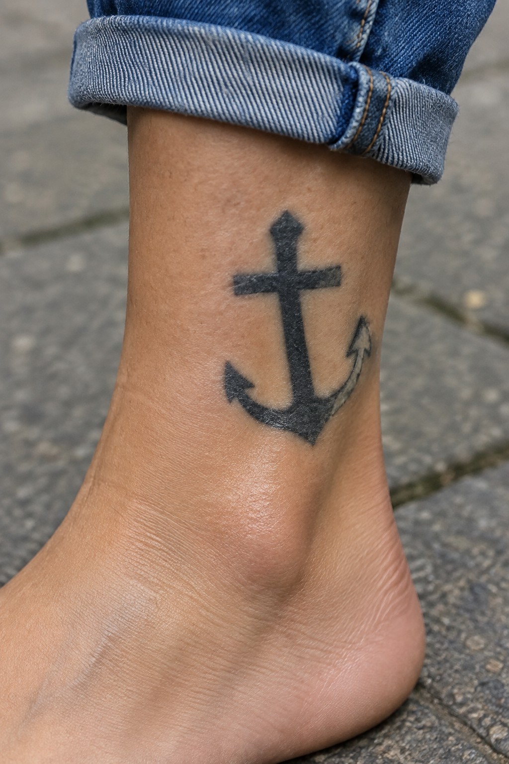 Black anchor tattoo on ankle below rolled blue jeans cuff on pavement