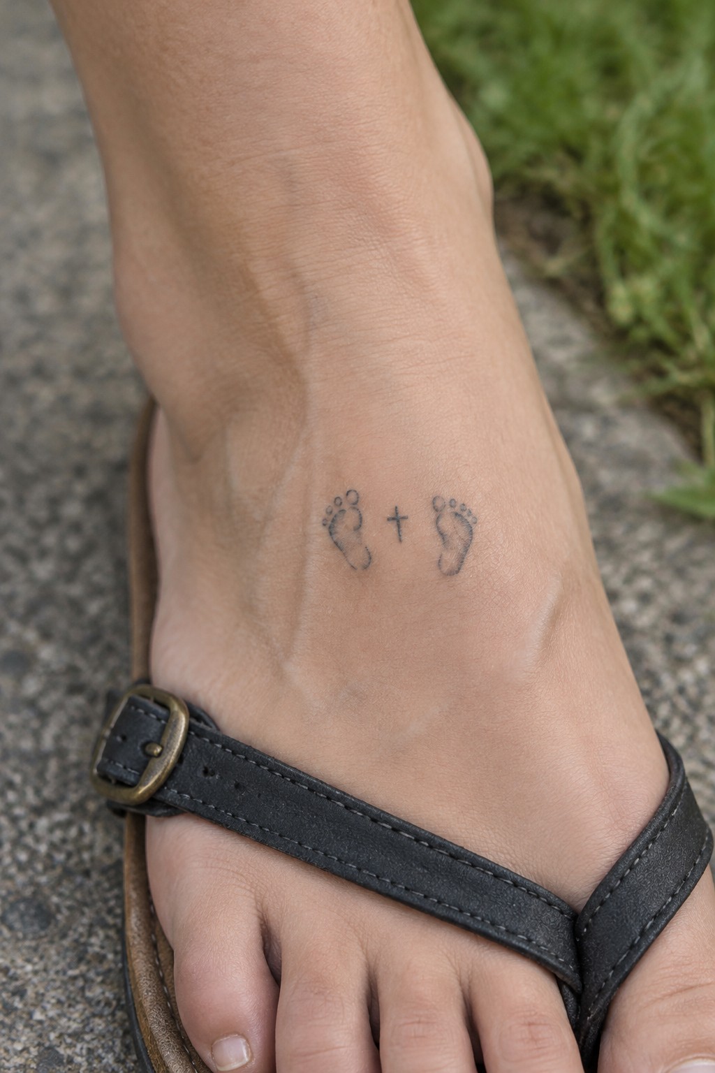 Black flip-flop sandal on foot with baby footprints and cross tattoo on ankle.