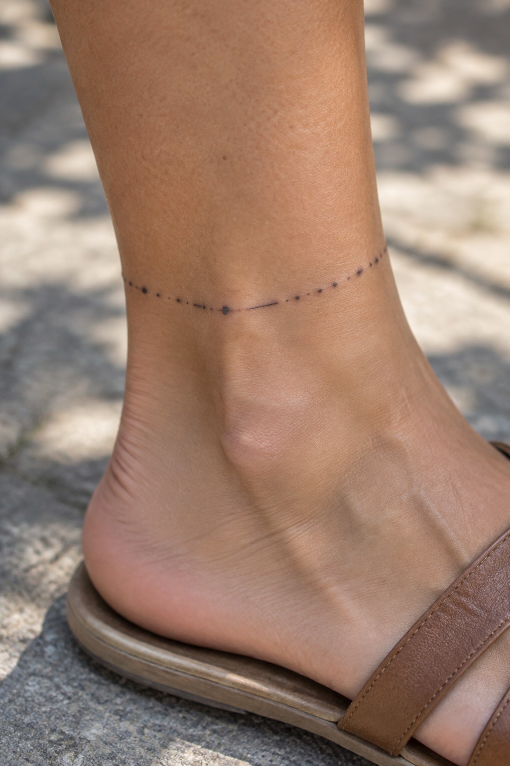 Ankle with black dotted line tattoo above brown leather sandal on pavement