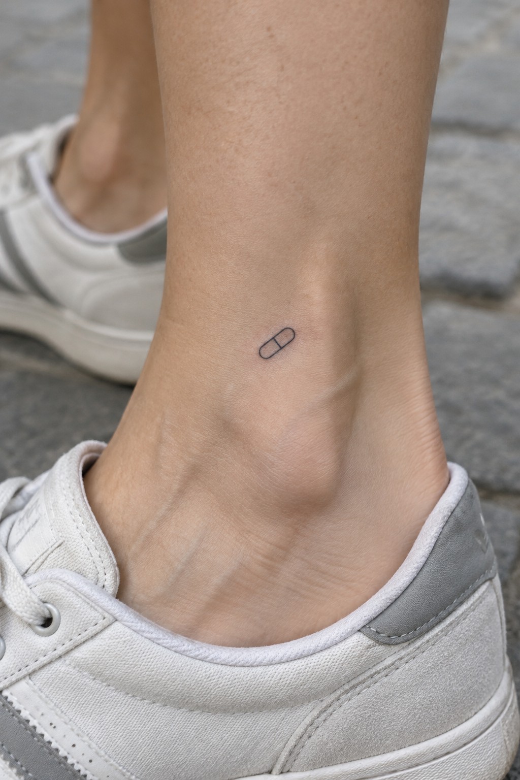 Small black capsule tattoo on ankle above white sneaker with gray accents.