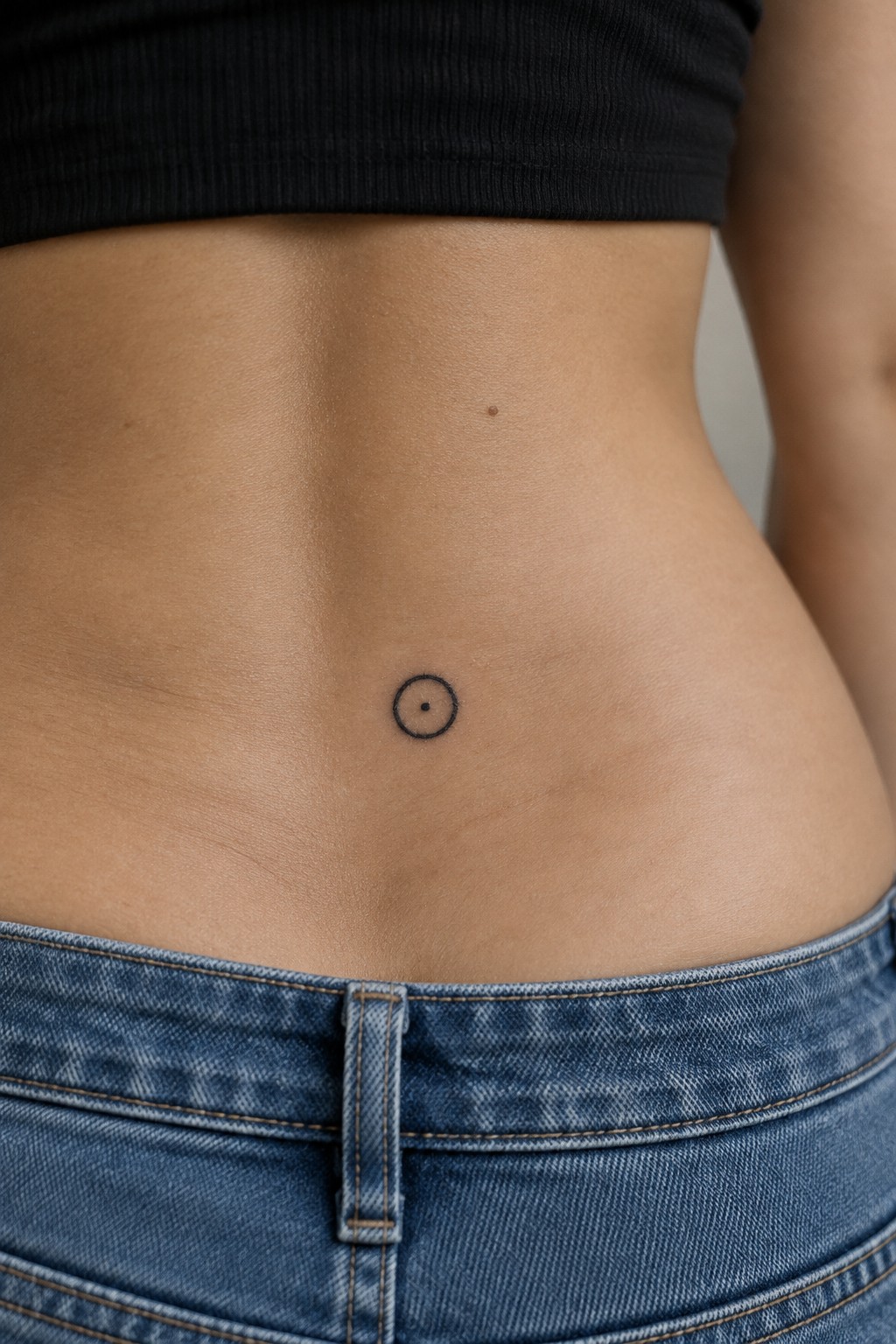 Bare midriff with small circular tattoo above blue denim jeans waistband.