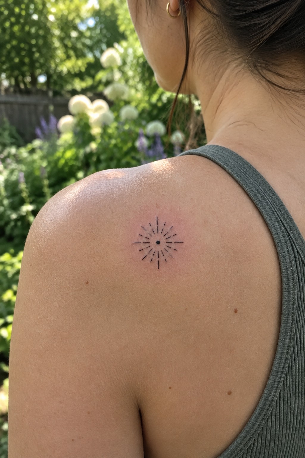 Black sunburst tattoo on shoulder above gray tank top in garden
