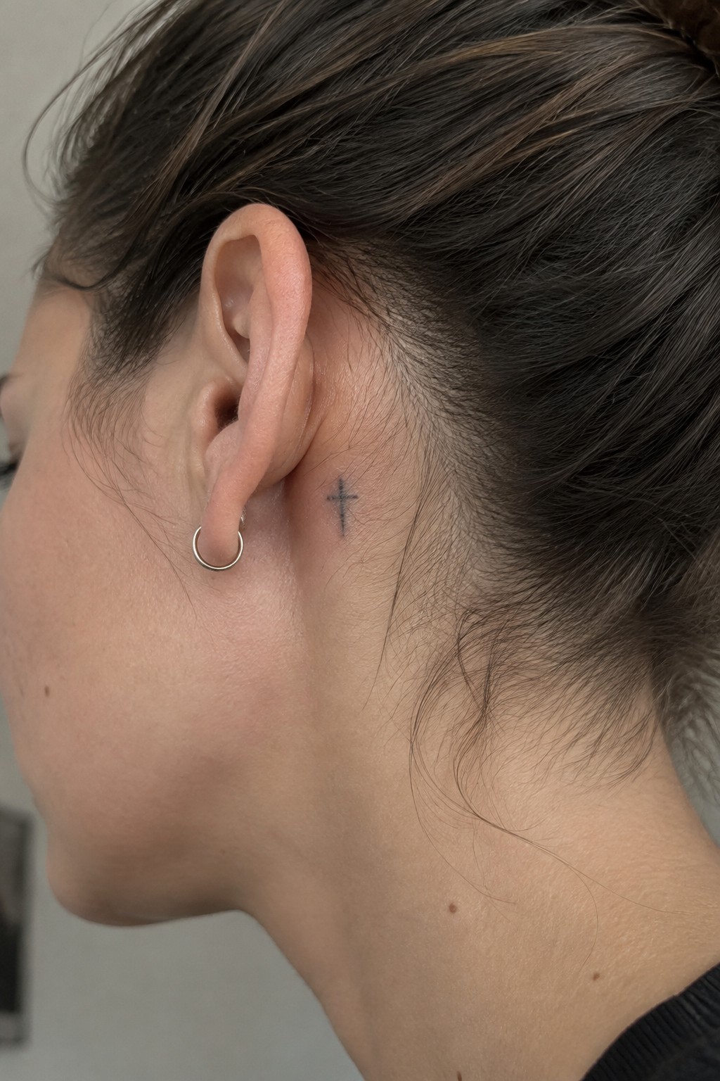 Small black cross tattoo placed behind the ear on the side of the head near the hairline