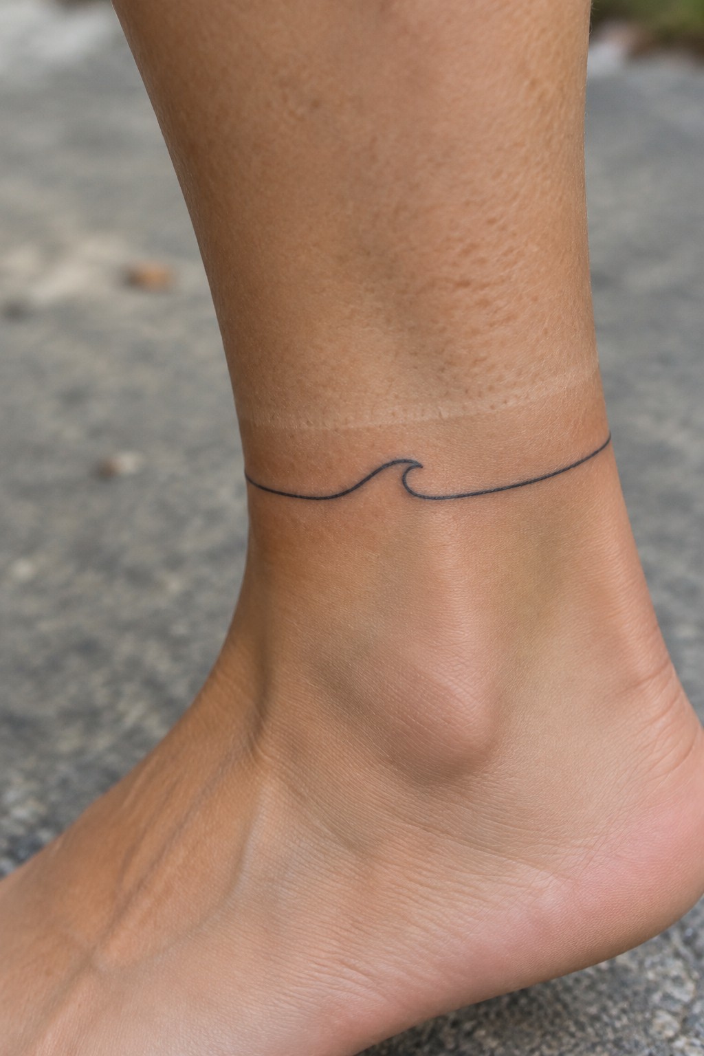 Fine-line black wave tattoo wrapping around a woman’s ankle.