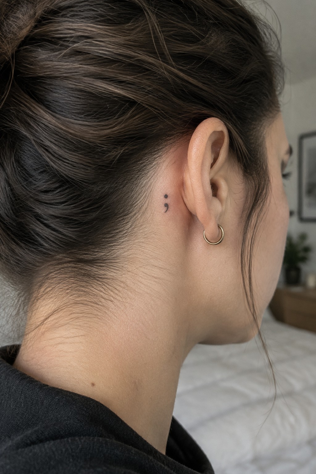 Small black semicolon tattoo behind a woman’s ear, above a gold hoop earring.