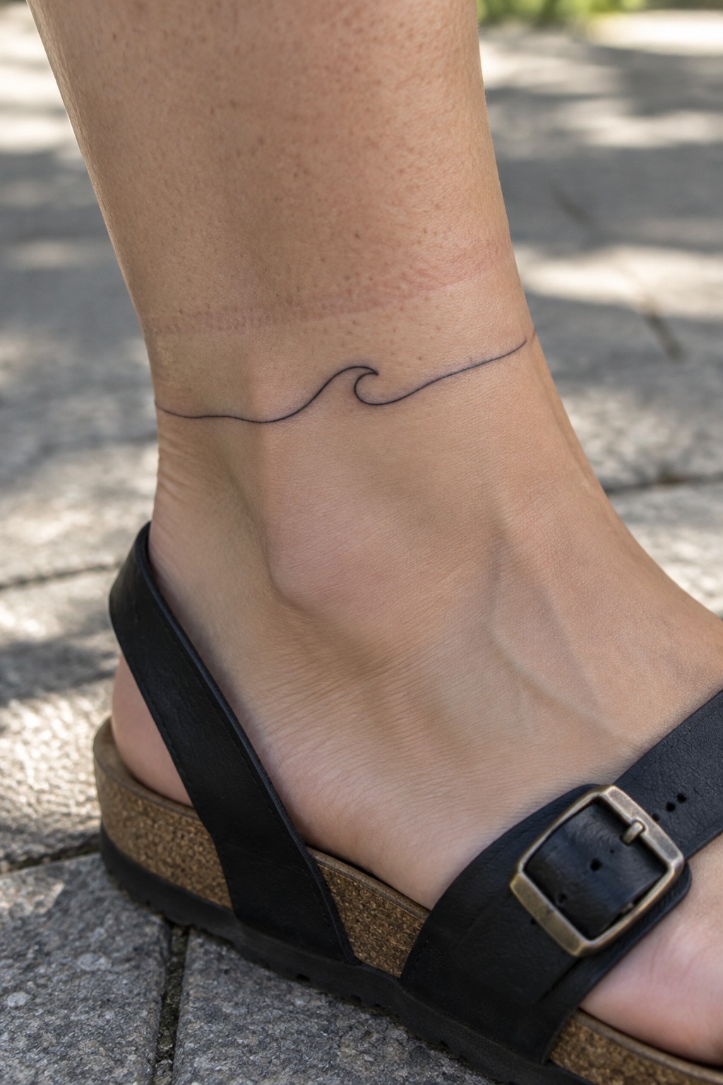 Small fine-line wave tattoo wrapping around the ankle above a black sandal.