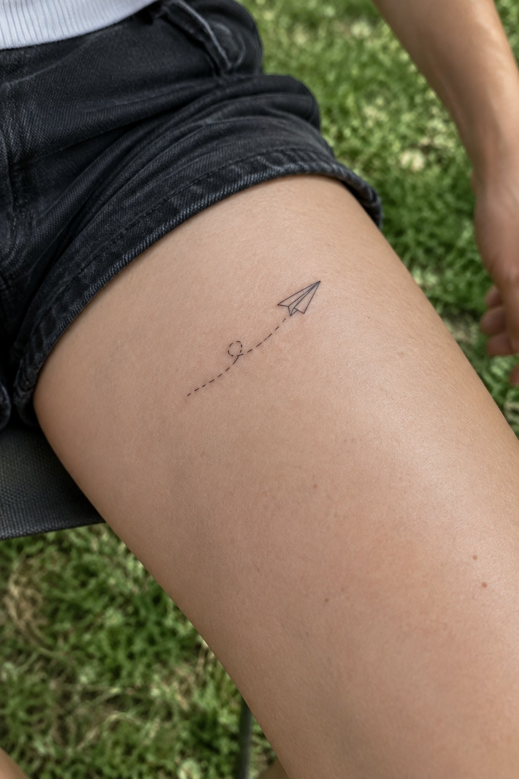Small black paper plane tattoo with a dotted curved flight path on the upper thigh.