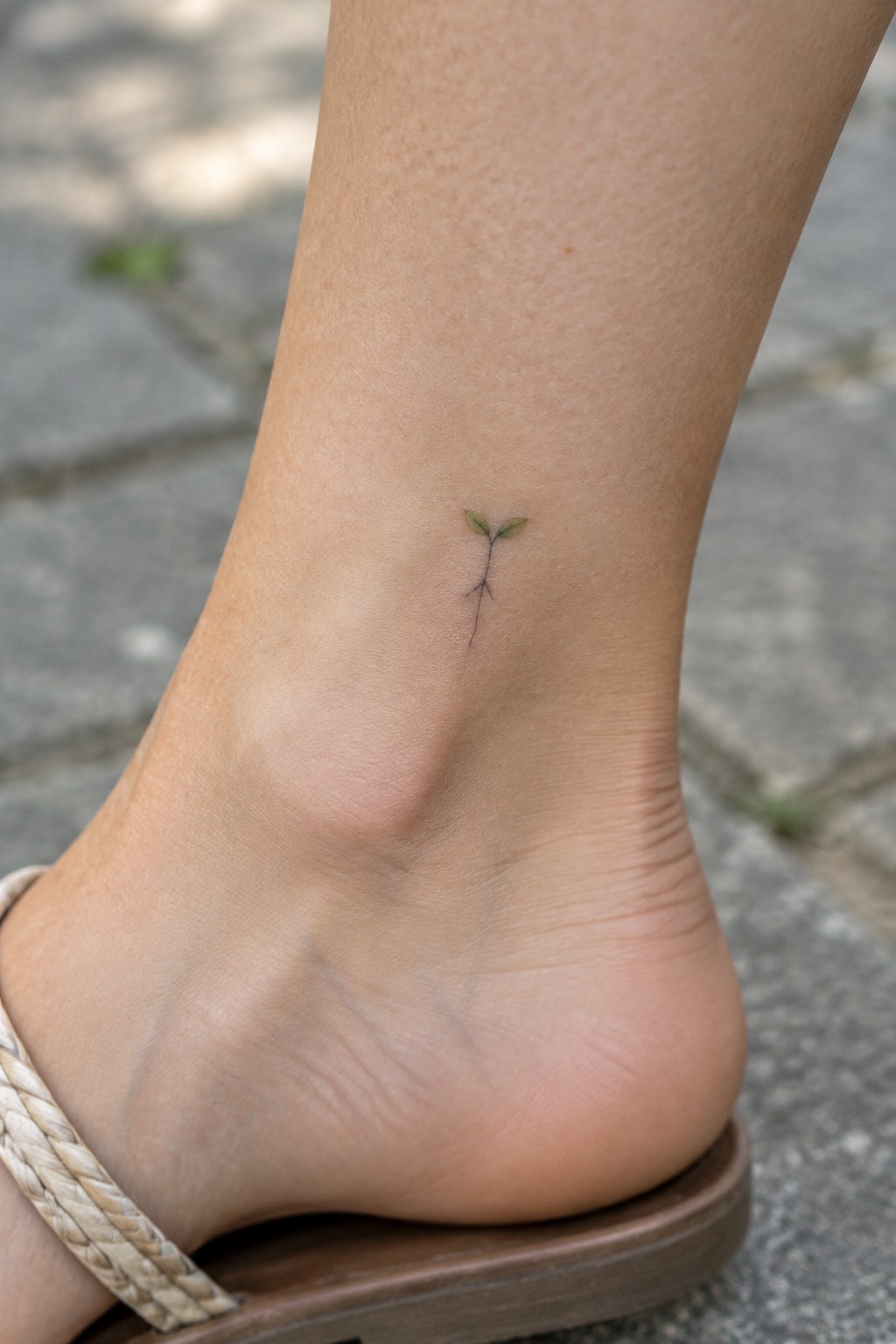 Tiny fine-line sprout tattoo with two green leaves on the inner ankle above a sandal.