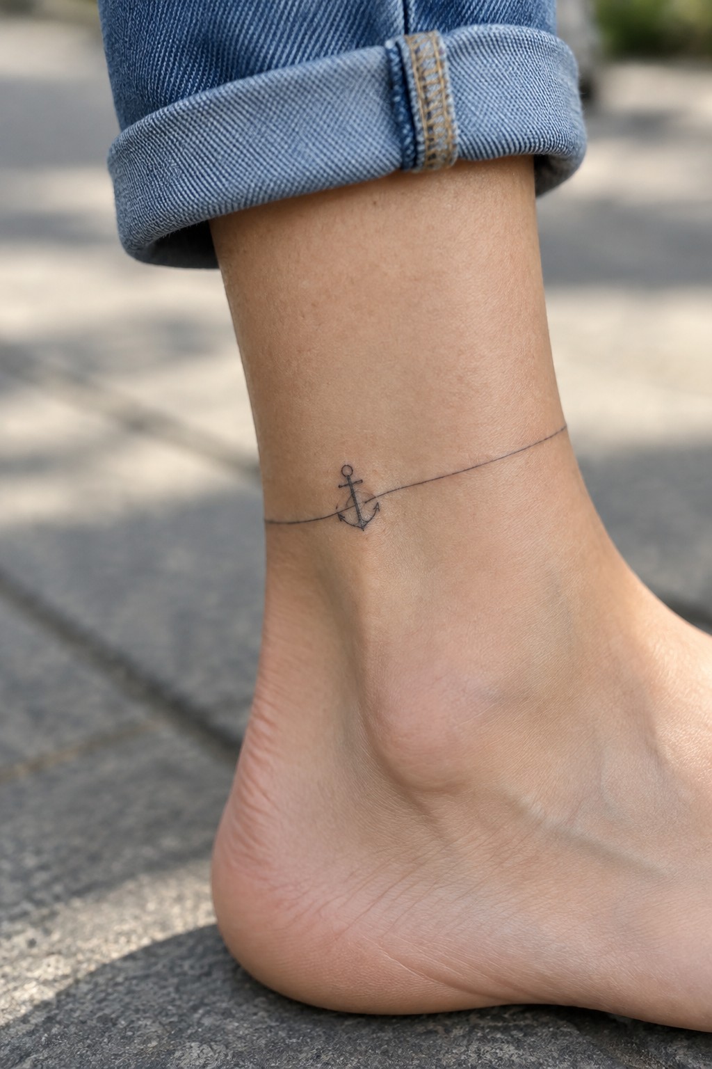 Small fine-line anchor tattoo on the ankle with a thin line wrapping around it, beneath a rolled-up denim cuff.