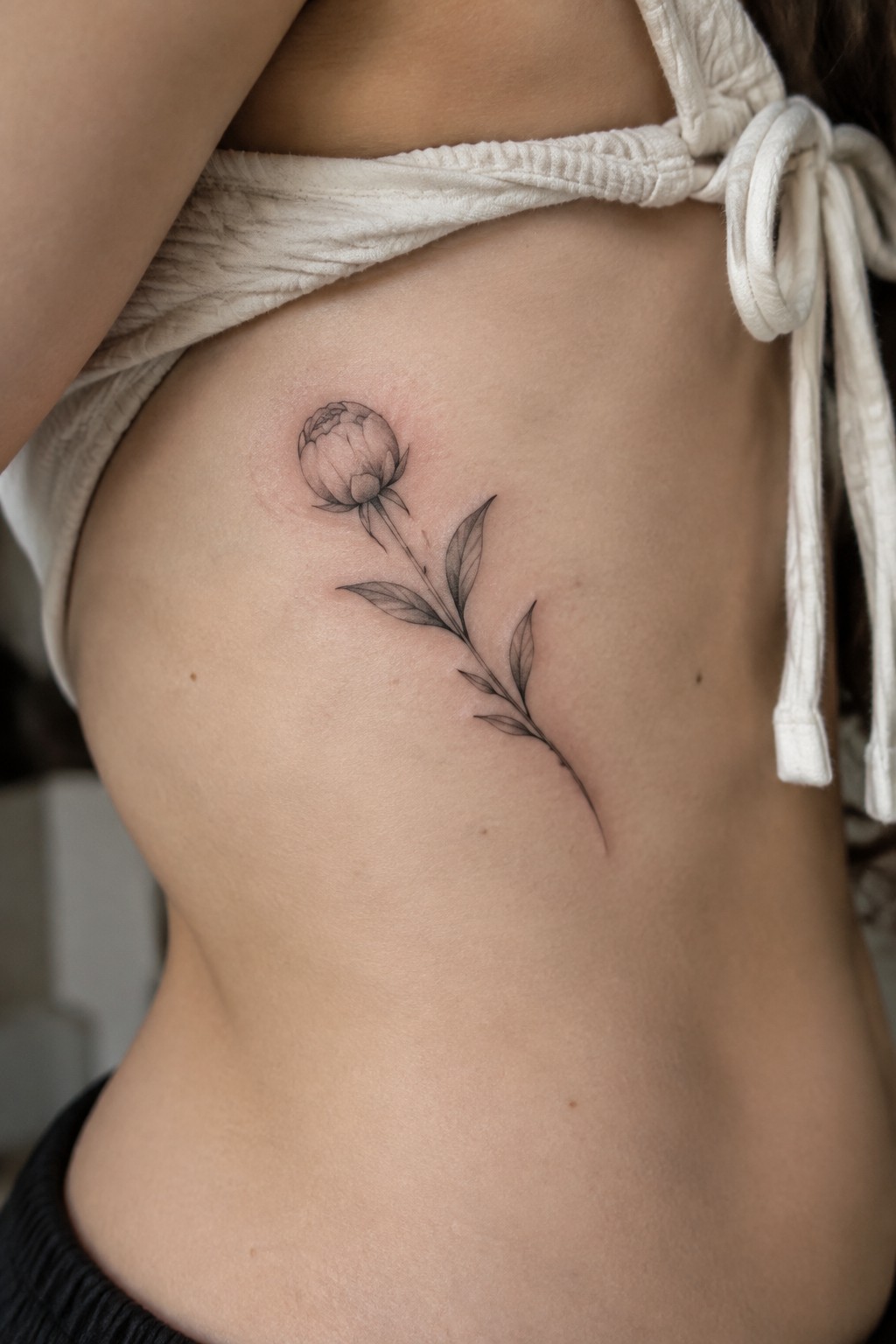 Fine-line black and gray flower tattoo with a slender leafy stem on a woman’s ribs.
