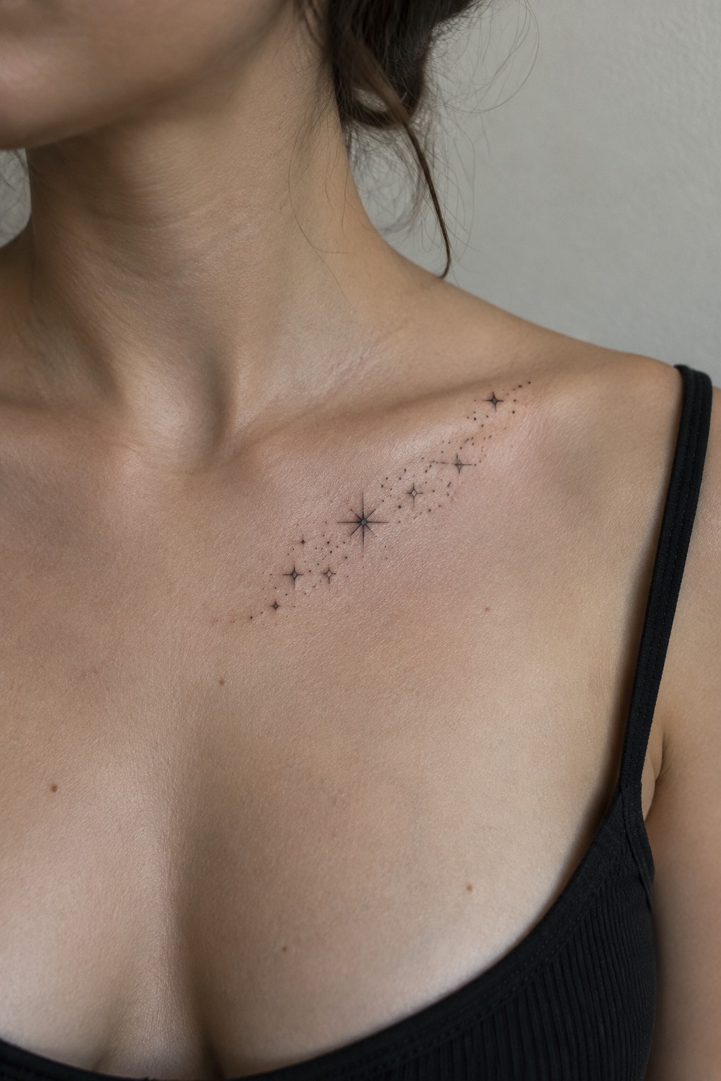 Fine-line constellation tattoo with small stars and dotted details running diagonally along a woman’s collarbone.