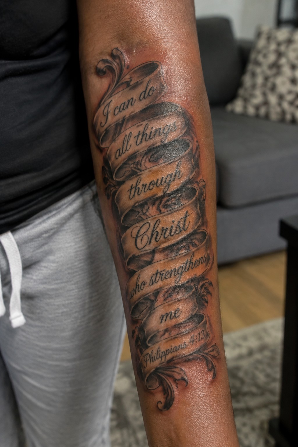 Black and gray ribbon tattoo on arm quoting Philippians 4:13 Bible verse