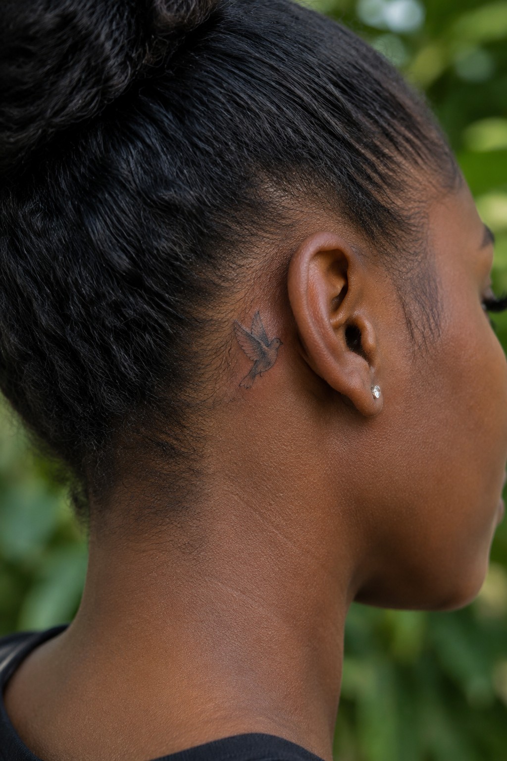 Dark hair in bun with bird tattoo behind ear and diamond stud earring.
