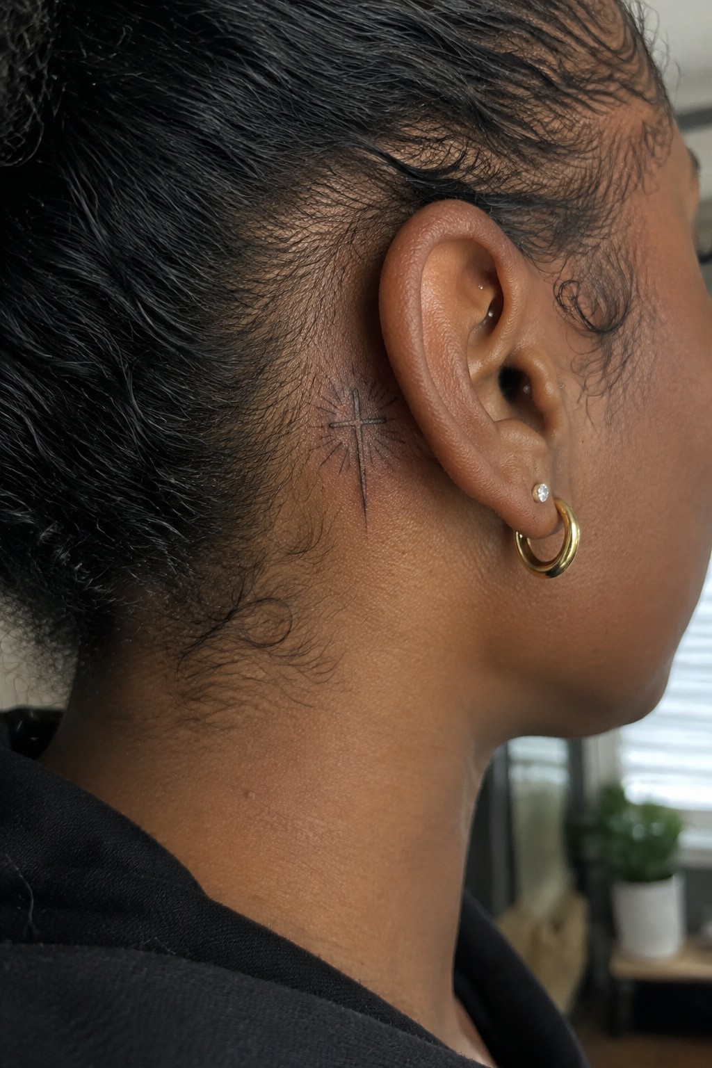 Small black and grey cross with rays tattooed behind the ear on dark skin