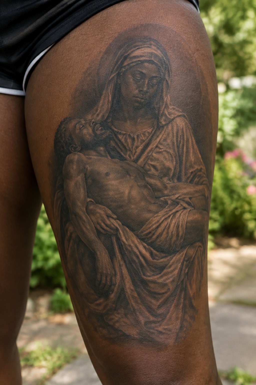 Black and grey pieta tattoo on a Black woman's upper thigh showing a robed woman cradling a man in her arms.